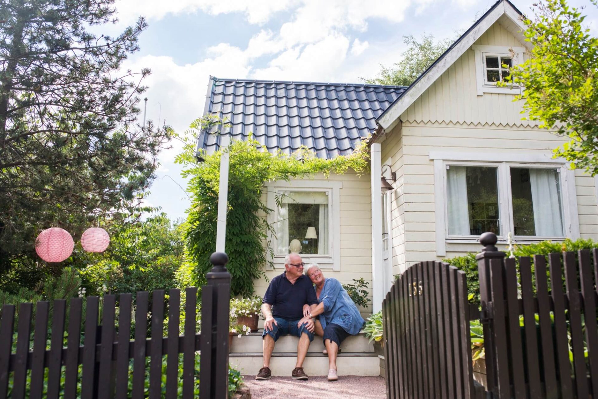 Mitt i en oas av grönt, står ett trähus med väggar målade i en ljus gul nyans och ett svart tak, hem till ett äldre par som sitter på dess veranda